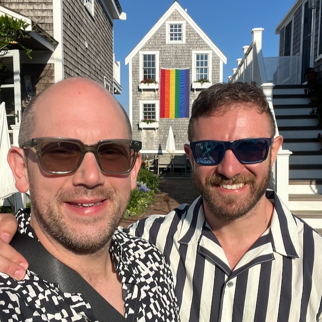 Elliot Milco and his ex on vacation together in Provincetown. We are standing next to each other and a house with a large pride flag is visible behind us.